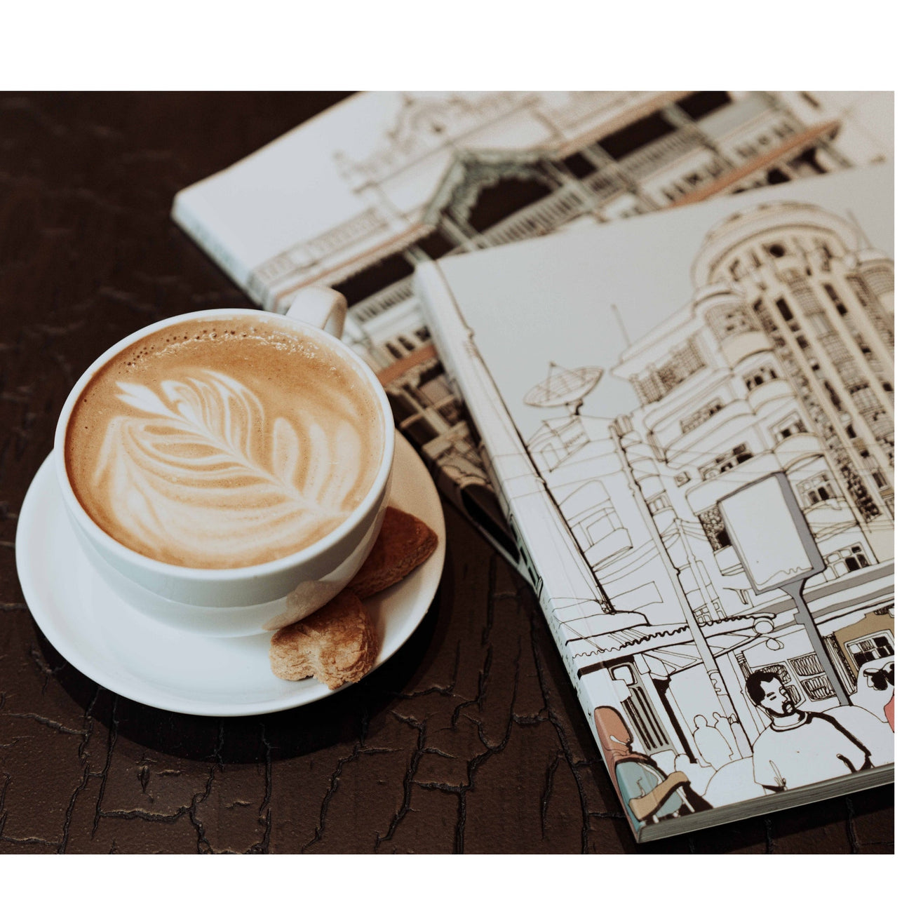 A latte with latte art and biscotti beside an open illustrated city journal on a dark wooden café table