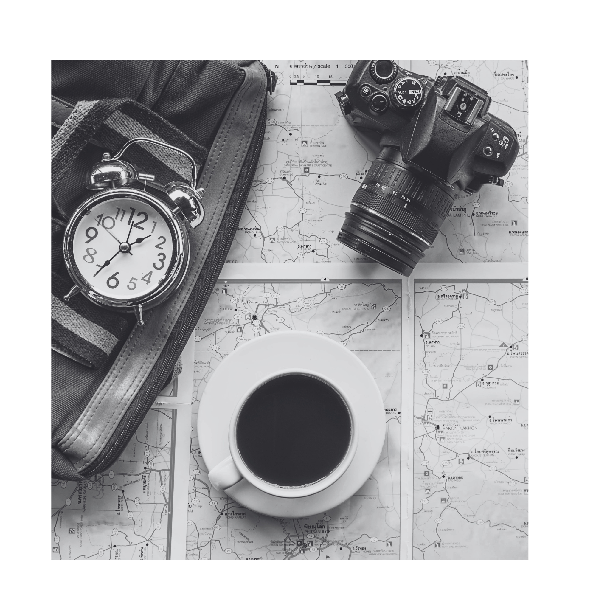Black and white overhead flat-lay of a travel bag, camera, alarm clock, road maps and a black coffee